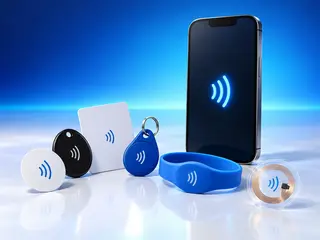 Un iPhone avec une icône NFC lumineuse derrière une rangée de formats de tags NFC : un autocollant, un jeton, une carte PVC, un porte-clés, un bracelet et un inlay transparent